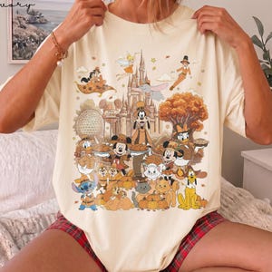 Puede incluir: Una camiseta de color crema con un gran estampado gráfico de varios personajes de Disney vestidos con ropa de temática otoñal. El diseño incluye un castillo, calabazas y follaje de otoño, con Mickey Mouse, Goofy y otros personajes.
