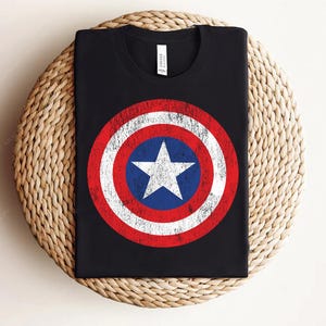 May include: Black t-shirt with a distressed shield graphic in red, white, and blue, with a white star at the centre. The design resembles a superhero's emblem. The tee is made of a soft material, suitable for casual wear and everyday use.