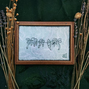 May include: A framed artwork featuring a line drawing of teeth hanging from a string, each adorned with a bow. The artwork is set against a light blue background with a textured appearance, within a brown frame. Dried flowers and foliage surround the frame.