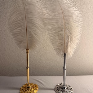 May include: Two decorative feather pens with white plumes. One pen has a gold-colored base and stem, while the other has a silver-colored base and stem. The bases are ornate and circular.
