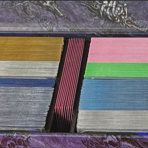 May include: A close-up of a box filled with stacks of colorful cards. The cards are arranged in rows of gold, silver, blue, pink, green, and red. The box has a decorative purple and black design.