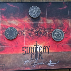 Könnte beinhalten: Eine rechteckige Spielmatte mit einem feurigen rot-schwarzen Hintergrund, dem Text SORCERY CON und einer Signatur. Drei kreisförmige Metall-Token mit komplexen geometrischen Gravuren liegen auf der Matte, die durch ein weißes Gitter unterteilt ist.