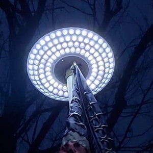 May include: A white LED light fixture with a circular design, mounted on a flagpole. The light fixture is surrounded by a ring of small, white LED lights.