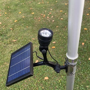 May include: Black solar-powered spotlight with a blue solar panel, mounted on a pole. The spotlight has a bright LED light and is attached to a black bracket. The background is green grass and a tree.