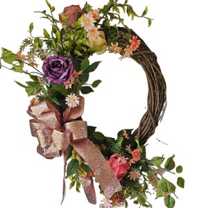 May include: A spring-themed wreath featuring a mix of pink, purple, and white flowers, greenery, and a rose gold glitter bow.