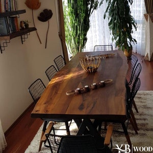 Puede incluir: Una gran mesa de comedor de madera con un diseño de borde vivo. La mesa está hecha de madera oscura y tiene un aspecto natural y rústico. Está rodeada de sillas de metal negro con un diseño de malla de alambre. La mesa está puesta con una cesta tejida y velas.