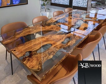 May include: A large, wooden dining table with a clear epoxy resin top. The table has a natural wood grain pattern and features a unique river table design. The table is surrounded by brown leather chairs.