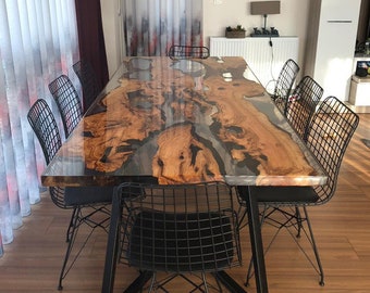 May include: A large wooden dining table with a clear epoxy resin top. The table has a natural wood grain pattern and is surrounded by black metal chairs.