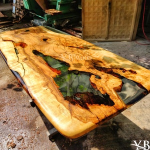 Op de afbeelding: Een groot, rechthoekig houten tafelblad met een natuurlijke houtnerf. Het tafelblad heeft een vulling van heldere epoxyhars met een zwart en bruin swirl-patroon. Het tafelblad staat op een metalen onderstel.
