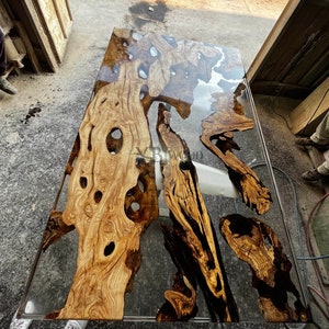 Op de afbeelding: Een groot, rechthoekig tafelblad gemaakt van natuurlijk hout met een heldere epoxyharsafwerking. Het hout heeft een natuurlijke nerf en verschillende knoesten. De hars is helder en glad en creëert een uniek en mooi effect.