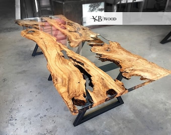 May include: A large wooden table with a clear epoxy resin top. The table has a natural wood grain and a unique shape. The table is supported by a black metal frame.
