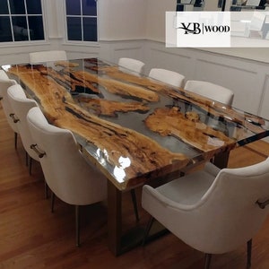 Op de afbeelding: Een grote, rechthoekige eettafel met een helder epoxyharsblad, met natuurlijke houtnerfpatronen. De tafel heeft een metalen onderstel en is omringd door wit gestoffeerde stoelen. De tafel is ongeveer 3 meter lang.