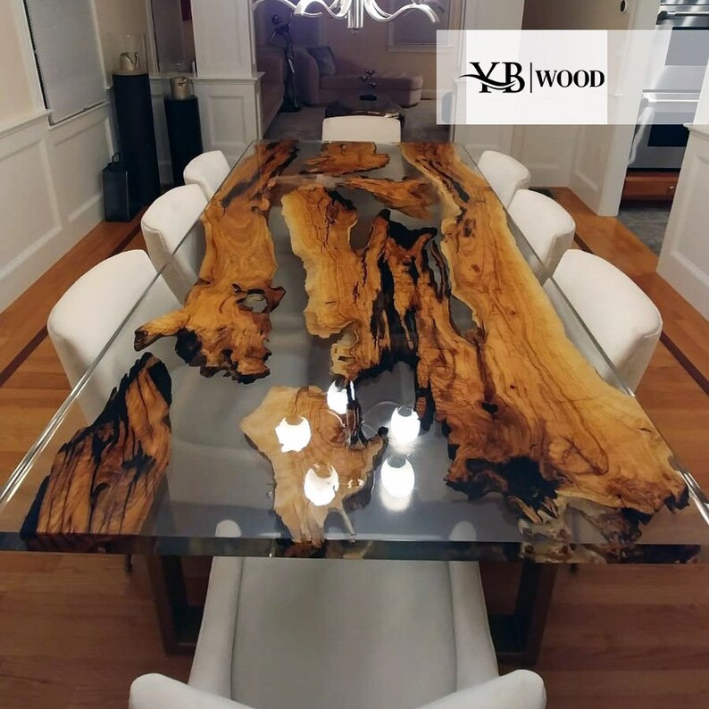Op de afbeelding: Een grote, rechthoekige eettafel met een helder harsblad, met natuurlijke houtnerfpatronen in bruin en zwart. De tafel wordt ondersteund door een donker metalen frame en is omgeven door witte stoelen. Het logo "YB WOOD" staat rechtsboven.
