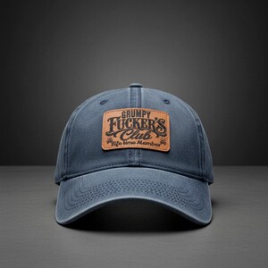 May include: A navy blue baseball cap featuring a rectangular tan leather patch centered on the front. The patch is embossed with the black text "GRUMPY FUCKER'S Club" and "life time Member" with small skull icons on the sides of the bottom line.