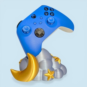 Puede incluir: Un mando azul de Xbox descansa sobre un soporte decorativo. El soporte presenta una luna creciente y estrellas doradas, con una base gris en forma de nube. El mando tiene botones negros y una cruceta gris.