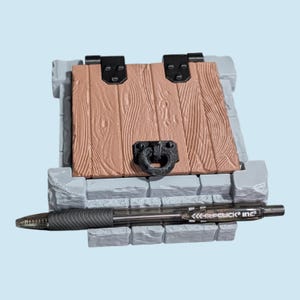 Puede incluir: Un modelo en miniatura de una puerta de madera con un tirador y bisagras negros, enmarcada en una estructura de piedra gris. Un bolígrafo negro y transparente descansa junto al modelo. La puerta es de color marrón claro con detalles de vetas de madera.
