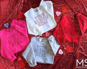 Puede incluir: Cuatro sudaderas de diferentes colores, rosa, blanco, rojo y gris, con el texto "YOUR DESIGN HERE" impreso en cada una. Las sudaderas están dispuestas sobre un fondo rojo brillante con caramelos en forma de corazón.