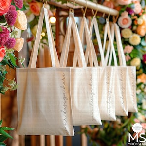 Pu&ograve; includere: Cinque borse tote in tela bianca appese a un rack di legno. Le borse hanno il testo "your design here" stampato sul lato. Le borse sono davanti a uno sfondo floreale.