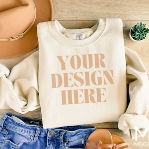 Puede incluir: Una sudadera de color crema con el texto "YOUR DESIGN HERE" impreso en color marr&oacute;n claro. La sudadera est&aacute; colocada sobre una superficie de madera con un sombrero marr&oacute;n, unos vaqueros azules y unas sandalias cerca.
