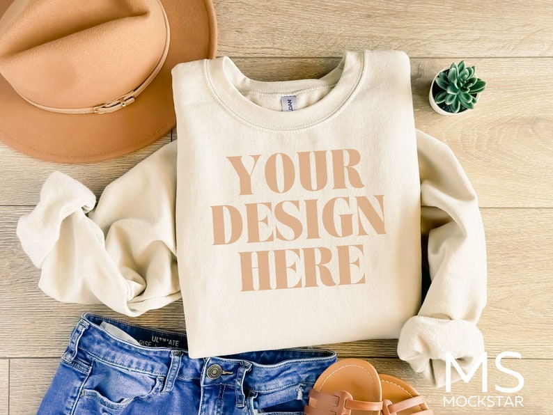 Puede incluir: Una sudadera de color crema con el texto "YOUR DESIGN HERE" impreso en color marr&oacute;n claro. La sudadera est&aacute; colocada sobre una superficie de madera con un sombrero marr&oacute;n, unos vaqueros azules y unas sandalias cerca.
