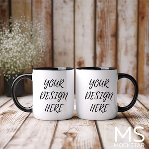 May include: Two white ceramic mugs with black rims and handles. The mugs are on a wooden surface and both have the text "YOUR DESIGN HERE" printed on them.