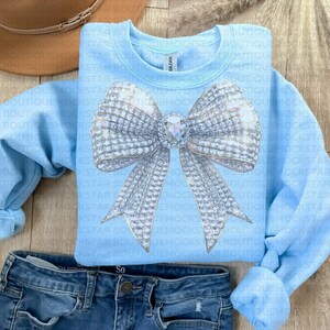 May include: A light blue sweatshirt with a large silver sparkly bow on the front. The bow has a large rhinestone in the center.