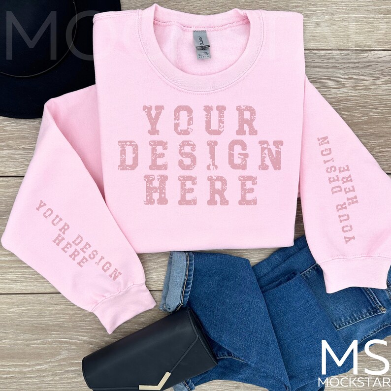 May include: Pink sweatshirt with the text "YOUR DESIGN HERE" printed on the front and sleeves. The sweatshirt is laid flat on a wooden surface with a pair of blue jeans and a black purse.