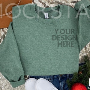 May include: A green crew neck sweatshirt with the text "YOUR DESIGN HERE" printed on the front. The sweatshirt is laying on a sparkly surface with a brown hat, white shoes, and a sprig of greenery.