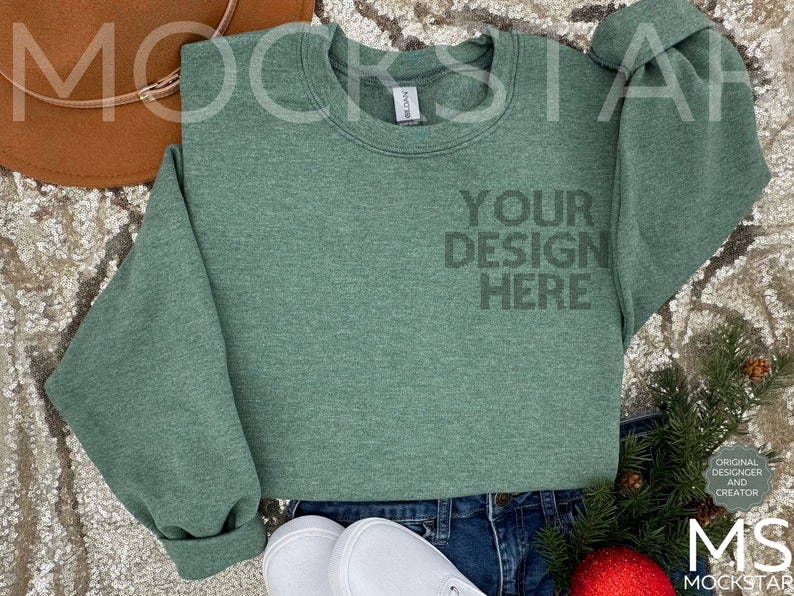 May include: A green crew neck sweatshirt with the text "YOUR DESIGN HERE" printed on the front. The sweatshirt is laying on a sparkly surface with a brown hat, white shoes, and a sprig of greenery.