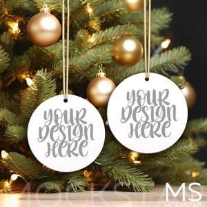 May include: Two white round ornaments with the text "Your design here" in gray lettering hanging on a Christmas tree with gold ornaments and white lights.