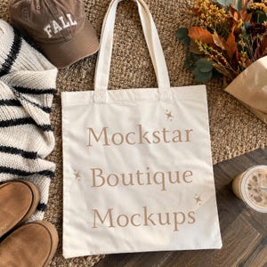 May include: A cream-colored canvas tote bag with two shoulder straps, featuring the text "Mockstar Boutique Mockups" printed in a light brown serif font, accented by small four-pointed star icons on the front surface of the bag.