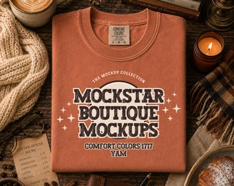 Könnte beinhalten: Ein orangefarbenes Comfort Colors T-Shirt mit dem Text "MOCKSTAR BOUTIQUE MOCKUPS" im Vintage-Stil. Das Shirt ist ordentlich auf einer Holzoberfläche gefaltet, umgeben von Kaffee, Büchern und einer Kerze.
