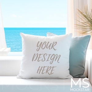 Puede incluir: Almohada blanca con el texto "YOUR DESIGN HERE" en un sofá blanco frente a una ventana con vista al mar.