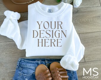 May include: White crew neck sweatshirt with the text "YOUR DESIGN HERE" printed in brown on the front. The sweatshirt is laid flat on a wooden surface with a pair of brown sandals and blue jeans.