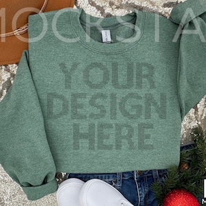 May include: A green heather sweatshirt with the text "YOUR DESIGN HERE" printed on the front. The sweatshirt is laying on a sparkly surface with a brown hat and a pair of white sneakers.