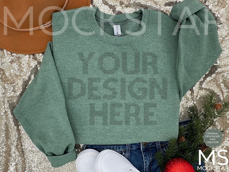 May include: A green heather sweatshirt with the text "YOUR DESIGN HERE" printed on the front. The sweatshirt is laying on a sparkly surface with a brown hat and a pair of white sneakers.