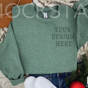 May include: A green sweatshirt with the text "YOUR DESIGN HERE" printed on the front. The sweatshirt is laying on a sparkly surface with a brown hat, jeans, and white shoes.