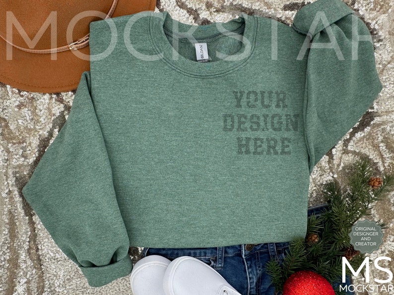 May include: A green sweatshirt with the text "YOUR DESIGN HERE" printed on the front. The sweatshirt is laying on a sparkly surface with a brown hat, jeans, and white shoes.