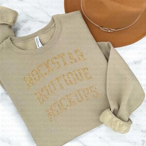 Pu&ograve; includere: Una felpa beige con il testo "Rockstar Boutique Mockups" in lettere di glitter dorate.