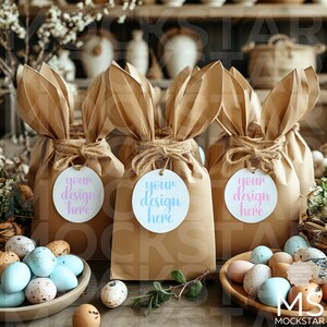 Op de afbeelding: Drie bruine papieren zakken met konijnenoren, vastgebonden met touw en versierd met ronde witte labels waarop in roze en blauwe letters "uw ontwerp hier" staat. De zakken zijn omringd door gevlekte paaseieren.