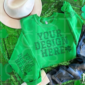 Puede incluir: Una sudadera verde con el texto "YOUR DESIGN HERE" y un gráfico de trébol. La sudadera se combina con unos vaqueros azules y un sombrero blanco. Recortes de tréboles verdes están dispersos alrededor de los artículos, todo sobre una superficie de lentejuelas verdes.