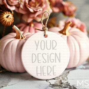 May include: A white ceramic ornament with the text "YOUR DESIGN HERE" in gray lettering. The ornament is hanging from a string and is sitting on a table with two pink ceramic pumpkins.