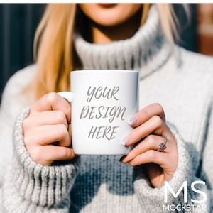 May include: White ceramic mug with the text "YOUR DESIGN HERE" in gray lettering. The mug is being held by a person wearing a gray sweater.