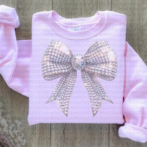 May include: A pink sweatshirt with a large sparkly silver bow design. The bow is made of rhinestones and has a gold center.