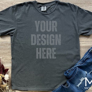 May include: A dark gray t-shirt with the text "YOUR DESIGN HERE" printed on the front. The shirt is paired with blue denim jeans. Dried flowers are arranged on the left side of the shirt.