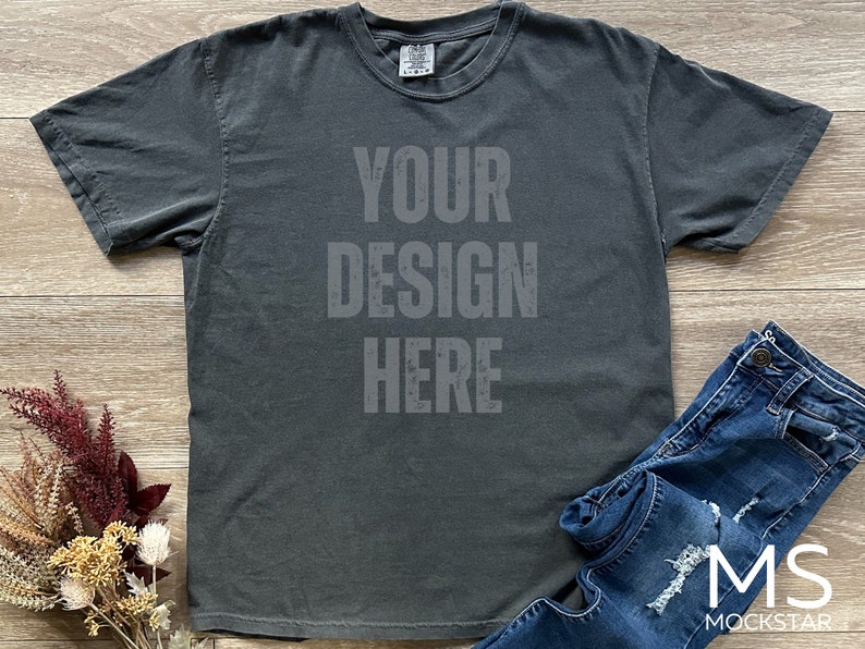 May include: A dark gray t-shirt with the text "YOUR DESIGN HERE" printed on the front. The shirt is paired with blue denim jeans. Dried flowers are arranged on the left side of the shirt.