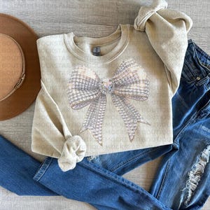 May include: A beige sweatshirt with a large sparkly silver bow design. The sweatshirt is laid out on a light wood surface with a brown hat and blue jeans.