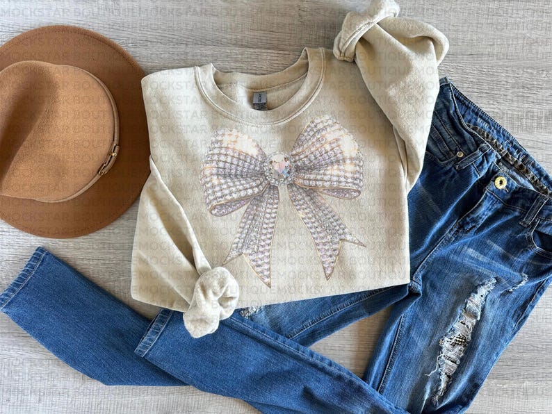 May include: A beige sweatshirt with a large sparkly silver bow design. The sweatshirt is laid out on a light wood surface with a brown hat and blue jeans.