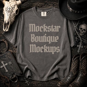 May include: A charcoal gray short-sleeve t-shirt with the text Mockstar Boutique Mockups printed in a light gray gothic font on the chest. The shirt features a crew neckline and a tag at the inner collar indicating a size large.