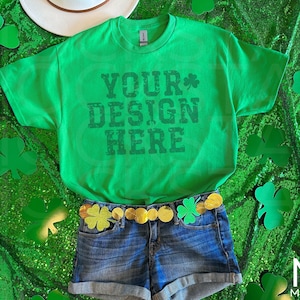 Puede incluir: Camiseta verde con el texto "YOUR DESIGN HERE" en una fuente desgastada. La camiseta está sobre una superficie verde brillante con recortes de trébol. Un par de pantalones cortos de mezclilla con un cinturón dorado y dijes de trébol es visible debajo de la camiseta.
